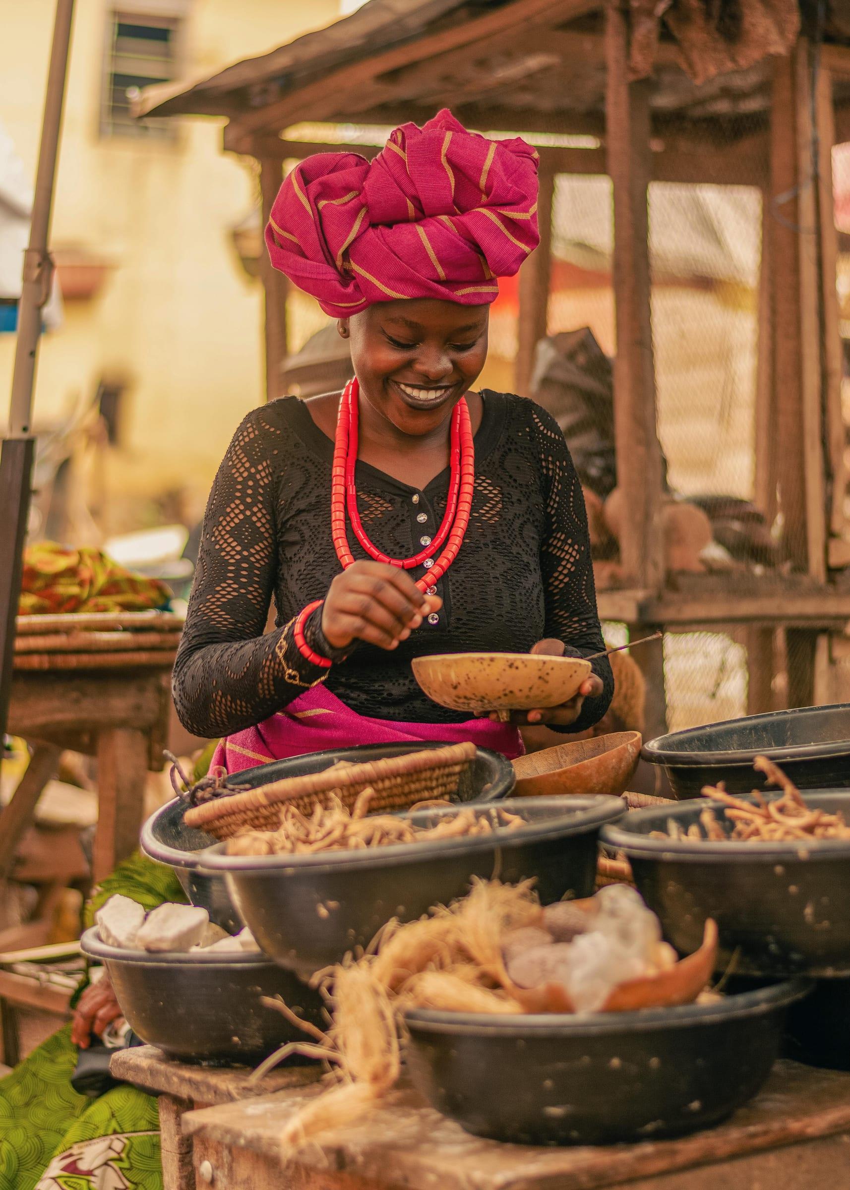 A market trader in a magenta head wrap and coral beads, smiling as she serves a portion from one of the bowls on her stall.