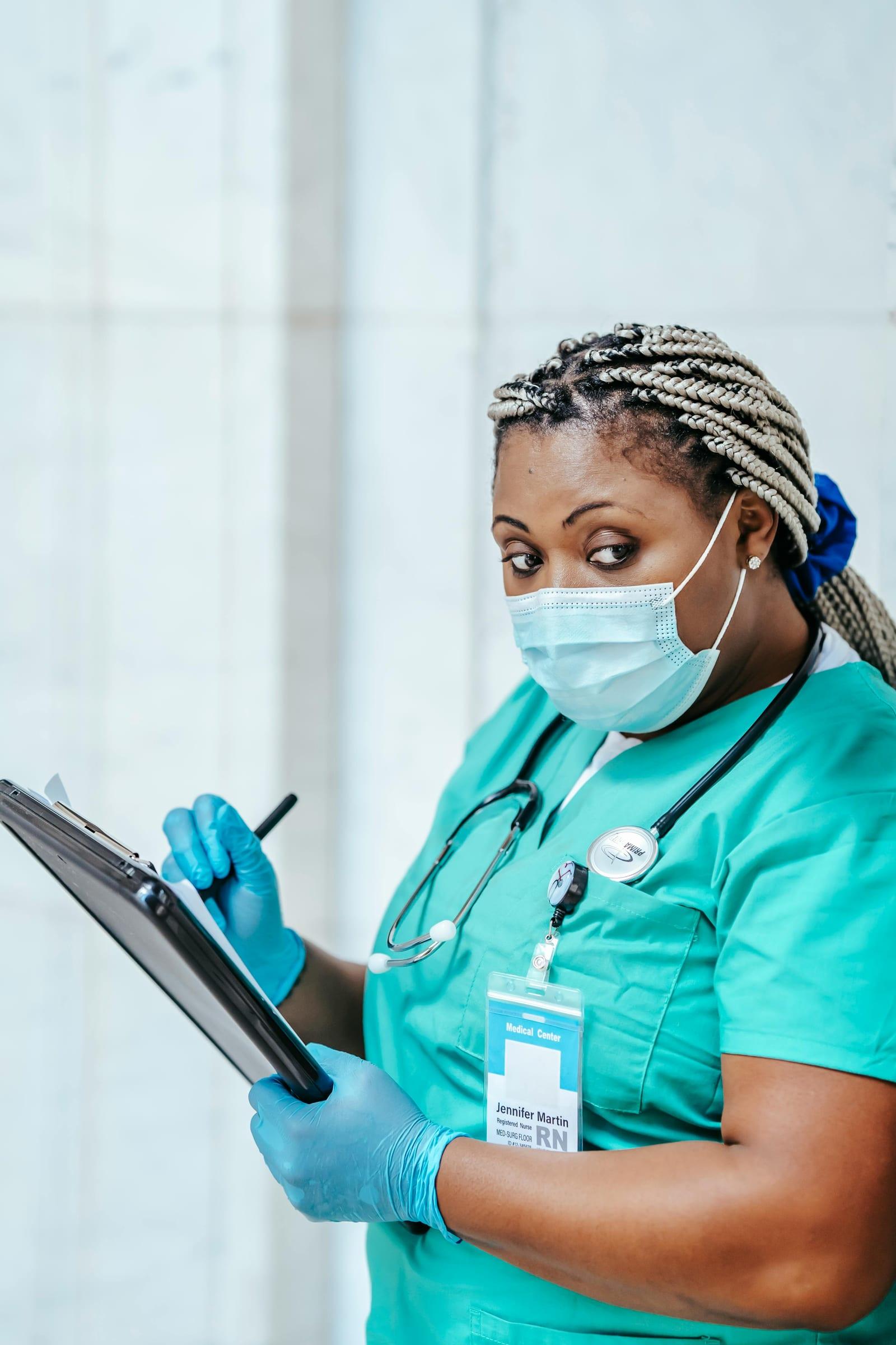 A nurse in green scrubs and a face mask, writing on a clipboard mid-shift.