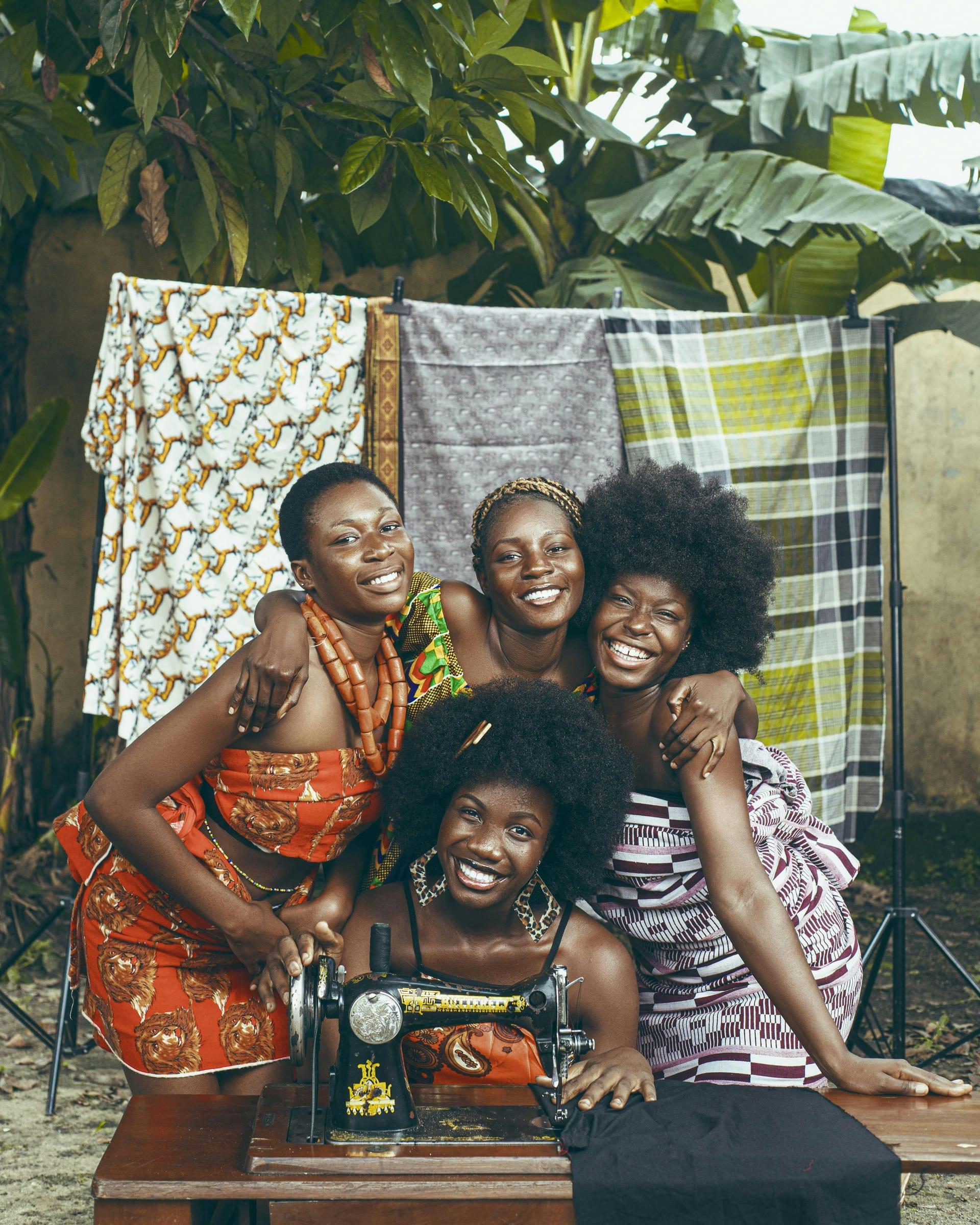 Four women gathered around a hand-crank sewing machine in a backyard, arms around each other, with patterned cloths hanging behind them.